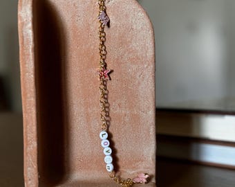 Könnte beinhalten: Eine goldene Kettenkette mit dem Wort "LOVER" in weißen Perlen. Die Kette hat rosa und weiße Stern- und Blumenanhänger.