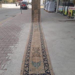 May include: An exceptionally long, narrow runner rug featuring a faded tan base color with intricate geometric and floral patterns in dark navy blue, brown, and muted teal. The rug has a distinct border and a repeating symmetrical design throughout.