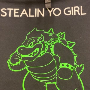 May include: Black t-shirt featuring the white text "STEALIN YO GIRL". Below the text is a green cartoon character. The shirt has a crew neck and a label.