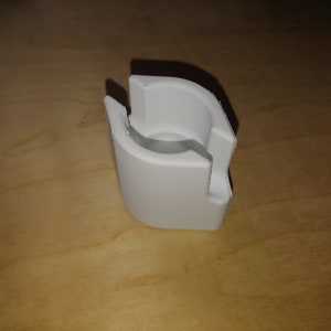 May include: A white plastic object with a curved design and a hollow center. The object has a rectangular shape with a cut-out section on one side. It is sitting on a light brown wooden surface.