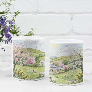 May include: Two white ceramic mugs with a rural landscape design. The design features a green hillside, blossom trees, sheep, and a stone cottage. The mugs have a white handle and rim.