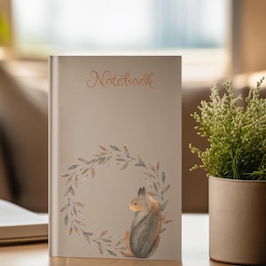 Könnte beinhalten: Ein beiges Notizbuch mit dem Wort "Notebook" in Schreibschrift. Das Cover zeigt eine Aquarellillustration eines Eichhörnchens in einem Blätterkranz. Das Notizbuch liegt auf einer Holzoberfläche.