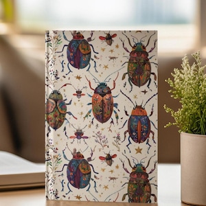 Puede incluir: Una libreta blanca con un patrón colorido de escarabajos y estrellas. Los escarabajos están dibujados en un estilo caprichoso con detalles intrincados y colores brillantes.