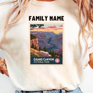 May include: Cream-colored t-shirt featuring a Grand Canyon National Park design. The design includes a landscape illustration within a postage stamp frame, with the text "FAMILY NAME" above and "GRAND CANYON NATIONAL PARK" below.