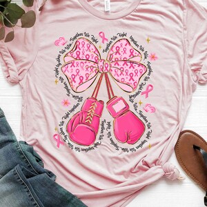 May include: Pink t-shirt featuring a large pink bow with pink ribbons and boxing gloves. The text "Together We Fight, Together We Win" is printed around the bow and gloves. The shirt is paired with blue jeans and sandals.