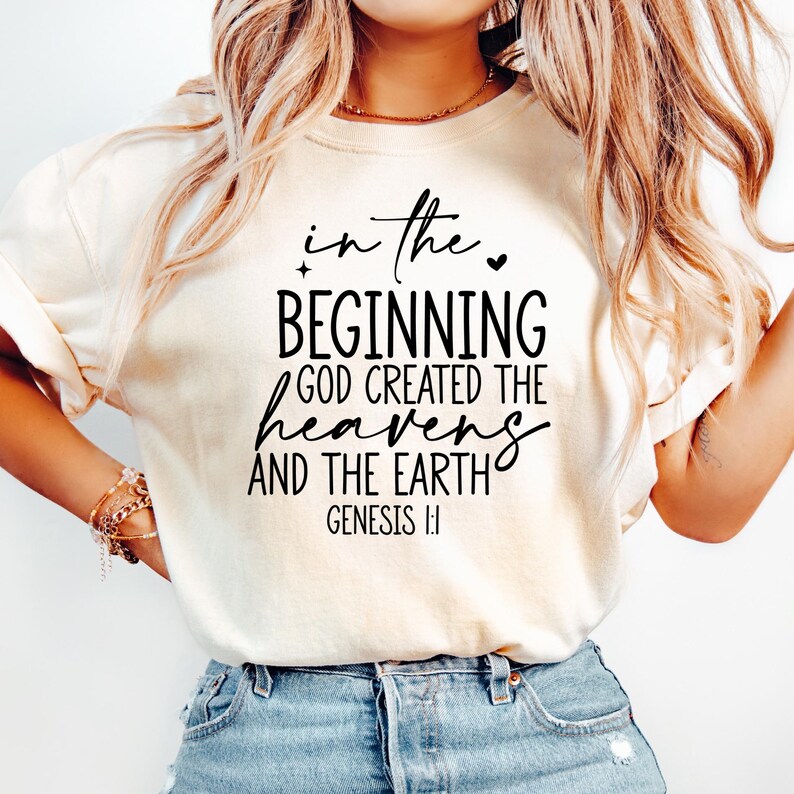 May include: Cream-colored t-shirt with the text "in the beginning God created the heavens and the earth" in black script. The text is from Genesis 1:1. The shirt is a casual style.