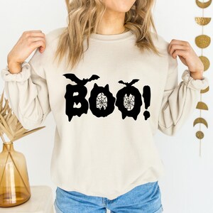 May include: A beige sweatshirt with the word "BOO!" in black, Halloween-themed lettering. The letters "O" are designed with spiderwebs, and bats are above the letters "B" and "O". The exclamation point is next to the last "O".