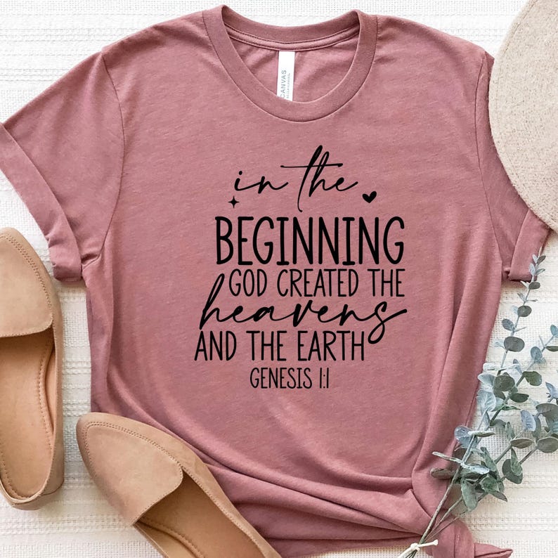 May include: A dusty rose t-shirt with the text "In the beginning God created the heavens and the earth" in black script. The shirt is paired with tan flats and a straw hat.