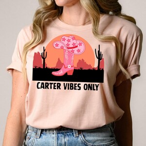 May include: A light pink t-shirt featuring a graphic of a pink cowboy boot and hat with star accents, set against a desert landscape with the text "CARTER VIBES ONLY".