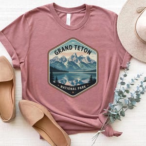 May include: Dusty rose t-shirt featuring a graphic of the Grand Teton National Park. The design includes a mountain range, lake, and the text "Grand Teton National Park". The shirt is paired with tan loafers and a straw hat.
