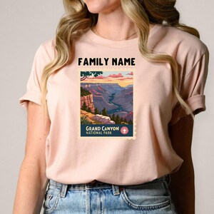 May include: A light pink t-shirt featuring a Grand Canyon National Park design. The design includes a landscape illustration with the text "FAMILY NAME" above and "GRAND CANYON NATIONAL PARK" below. The shirt is made of soft material.