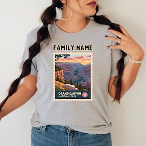 May include: A light gray t-shirt featuring a Grand Canyon National Park design. The graphic resembles a vintage postage stamp with a scenic canyon view, the text "FAMILY NAME" at the top, and "GRAND CANYON NATIONAL PARK" at the bottom.