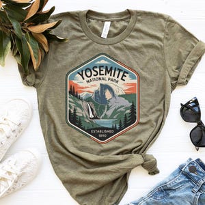 May include: Olive green t-shirt featuring a Yosemite National Park graphic. The graphic is a colorful illustration of a mountain landscape with the text "YOSEMITE NATIONAL PARK ESTABLISHED 1890". The shirt has rolled-up sleeves.