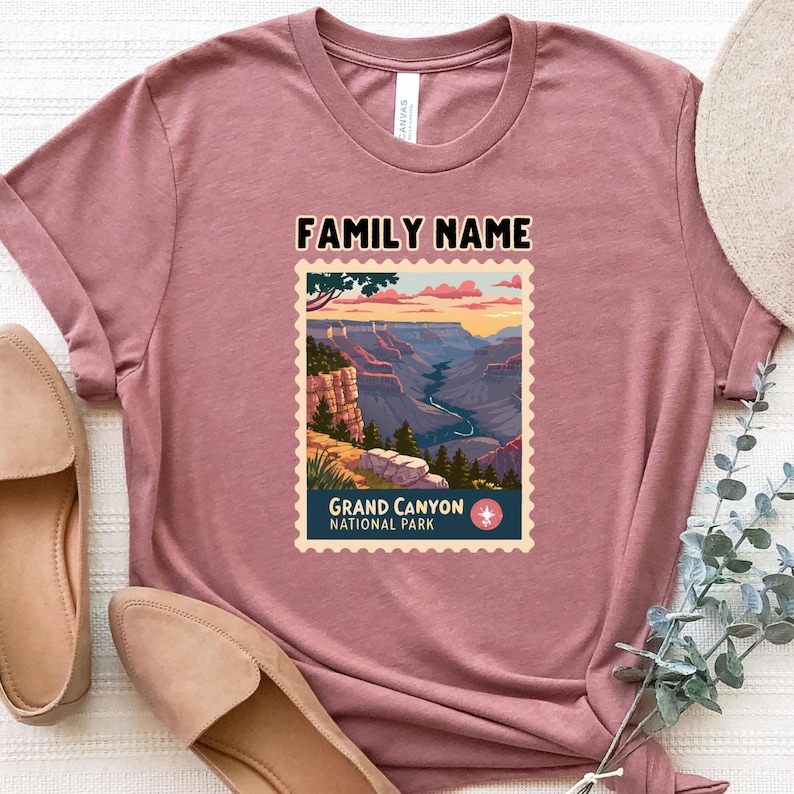 May include: Dusty rose t-shirt featuring a Grand Canyon National Park stamp design. The stamp includes a scenic canyon view with the text "FAMILY NAME" above and "GRAND CANYON NATIONAL PARK" below. The shirt is paired with tan flats.