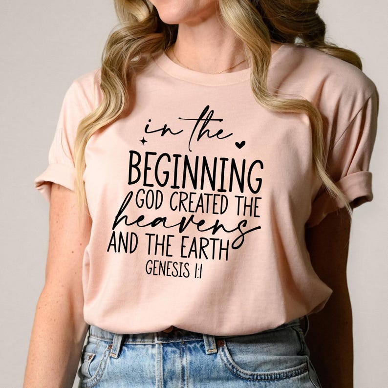 May include: A light pink t-shirt with rolled-up sleeves. The front of the shirt features black script that reads "In the beginning God created the heavens and the earth Genesis 1:1."