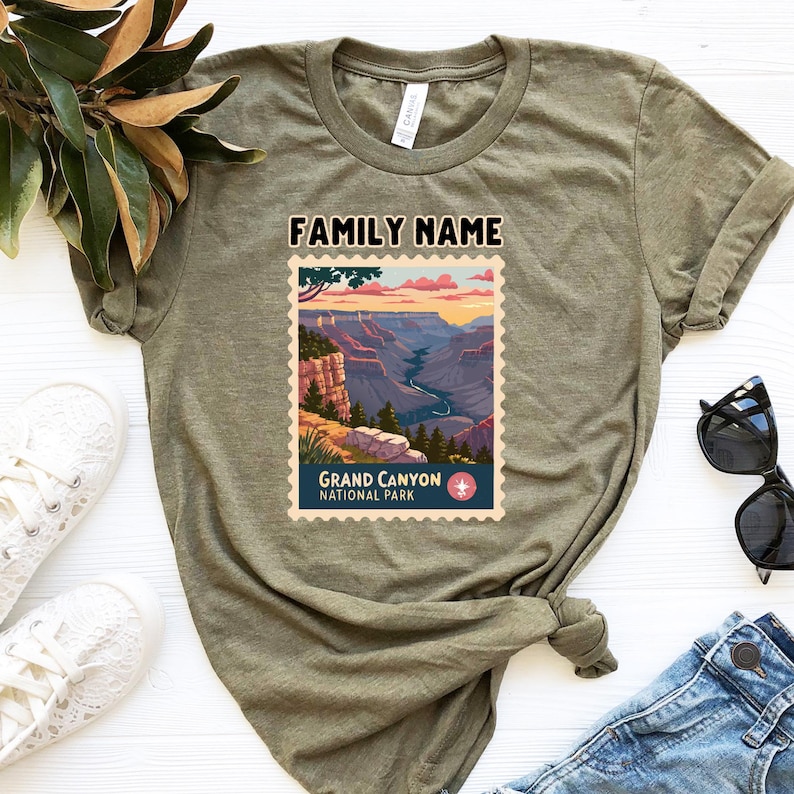 May include: Olive green t-shirt featuring a Grand Canyon National Park design. The graphic includes a scenic view of the canyon with the text "FAMILY NAME" above and "GRAND CANYON NATIONAL PARK" below. The shirt is paired with white sneakers and blue jeans.