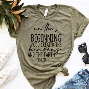 May include: Olive green t-shirt with the text "in the beginning God created the heavens and the earth" in black script. The shirt is paired with blue jeans, white sneakers, and sunglasses.