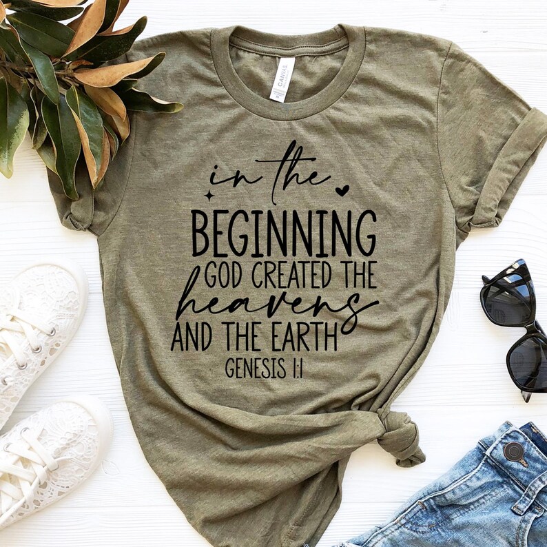 May include: Olive green t-shirt with the text "in the beginning God created the heavens and the earth" in black script. The shirt is paired with blue jeans, white sneakers, and sunglasses.