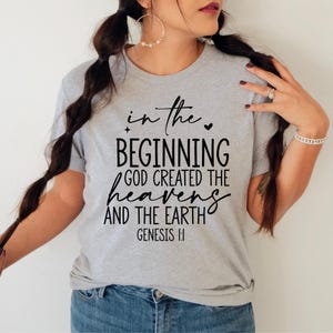 May include: A light gray t-shirt with the text "in the beginning GOD CREATED THE heavens AND THE EARTH Genesis 1:1" in black script. The shirt is a crew neck style.