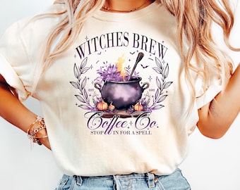 Può includere: T-shirt color crema con una grafica circolare raffigurante un calderone di strega, fiori viola e zucche. Il testo recita "Witches Brew Coffee & Co. Stop in for a Spell."