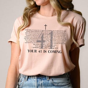 May include: A light pink t-shirt with black text and a cross graphic. The text includes the phrase "YOUR 41 IS COMING." and a passage about the Bible. The shirt is made of a soft material.