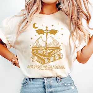 May include: Cream-colored sweatshirt with a gold graphic of a stack of books, a dragon, a sword, a crescent moon, and stars. The text "I AM THE SKY AND THE POWER OF EVERY STORM THAT HAS EVER BEEN. I AM INFINITE." is printed below the graphic.