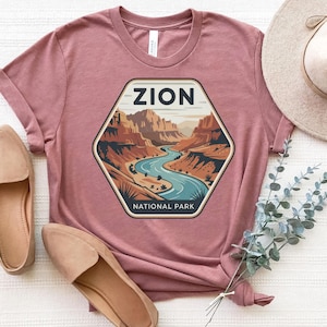 May include: A dusty rose t-shirt featuring a hexagonal graphic of Zion National Park. The graphic shows a river winding through a canyon, with the word "ZION" at the top and "NATIONAL PARK" at the bottom. The shirt is paired with tan flats and a straw hat.