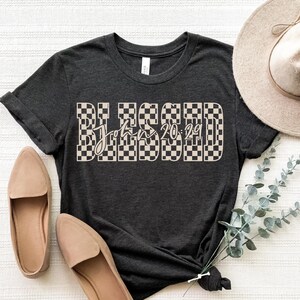 May include: Dark grey t-shirt featuring the word "BLESSED" in a checkered design, with "John 20:29" in script. The shirt is styled with tan loafers and a beige hat. A sprig of greenery is tied to the shirt.