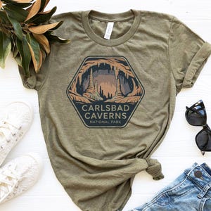 May include: An olive green t-shirt with a Carlsbad Caverns National Park graphic. The design is a hexagon with a cave illustration and the text "Carlsbad Caverns National Park". The shirt is styled with white trainers and blue jeans.