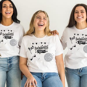 May include: Three women wearing white t-shirts with a black graphic that says "Bachelorette Party". The design includes a masquerade mask, martini glasses, and a disco ball. They are wearing blue jeans.