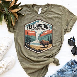 May include: Olive green t-shirt featuring a Yellowstone National Park graphic. The graphic includes a bison, geyser, and trees, with the text "YELLOWSTONE NATIONAL PARK". The shirt is paired with blue jeans, white sneakers, and sunglasses.