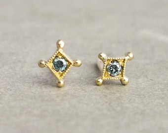 May include: A pair of gold earrings with a blue gemstone in the center of each earring. The earrings are in the shape of a square with rounded corners.