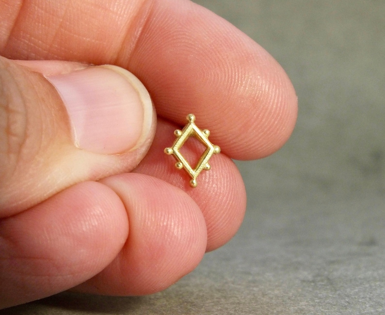 May include: A small gold-colored metal charm in the shape of a diamond with a square opening in the center. The diamond is surrounded by small circles.