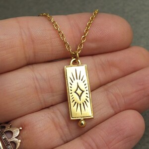 Może przedstawiać: Złoty naszyjnik z prostokątnym wisiorem w kształcie rozgwieżdżonego słońca. Wisiorek jest przymocowany do delikatnego złotego łańcuszka.