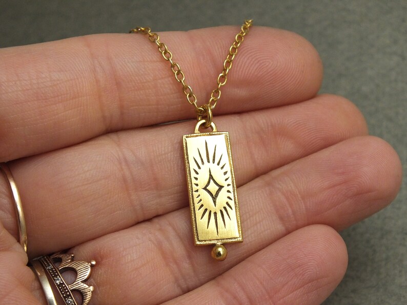 Może przedstawiać: Złoty naszyjnik z prostokątnym wisiorem w kształcie rozgwieżdżonego słońca. Wisiorek jest przymocowany do delikatnego złotego łańcuszka.