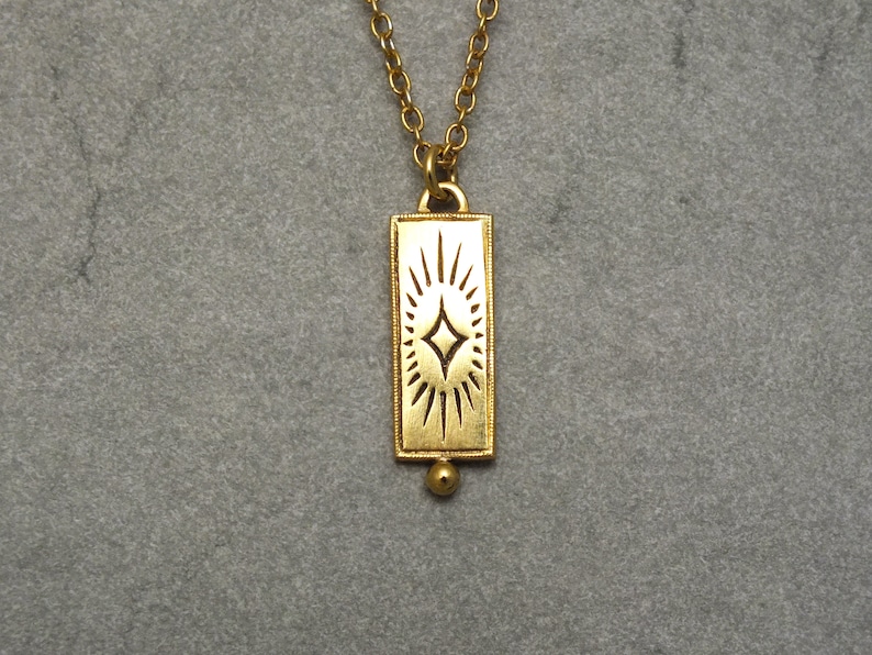 Może przedstawiać: Złoty naszyjnik z prostokątnym wisiorem w kształcie rozgwieżdżonego słońca. Wisiorek jest przymocowany do delikatnego łańcuszka.