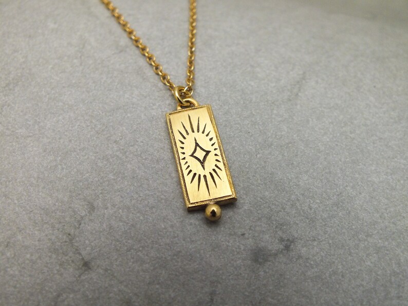 Może przedstawiać: Złoty naszyjnik z prostokątnym wisiorem przedstawiającym stylizowany wz&oacute;r gwiazdy.