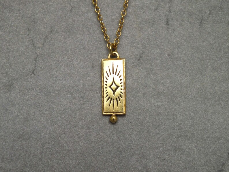 Może przedstawiać: Złoty naszyjnik z prostokątnym wisiorem przedstawiającym stylizowany wz&oacute;r gwiazdy.