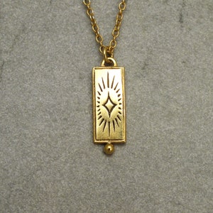 Może przedstawiać: Złoty naszyjnik z prostokątnym wisiorem w kształcie rozgwieżdżonego słońca. Wisiorek jest przymocowany do delikatnego złotego łańcuszka.