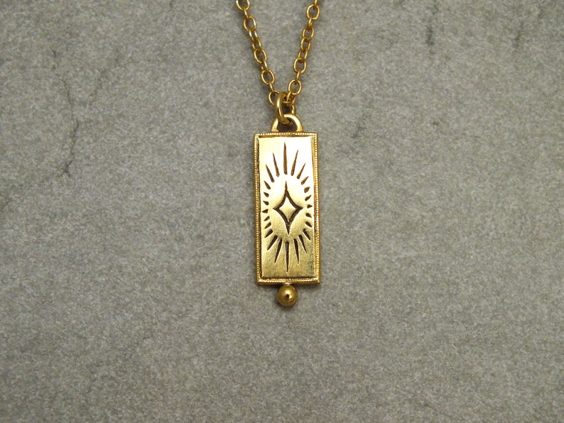 Może przedstawiać: Złoty naszyjnik z prostokątnym wisiorem w kształcie rozgwieżdżonego słońca. Wisiorek jest przymocowany do delikatnego złotego łańcuszka.