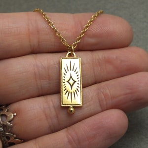 Może przedstawiać: Złoty naszyjnik z prostokątnym wisiorem w kształcie rozgwieżdżonego słońca. Wisiorek jest przymocowany do delikatnego złotego łańcuszka.