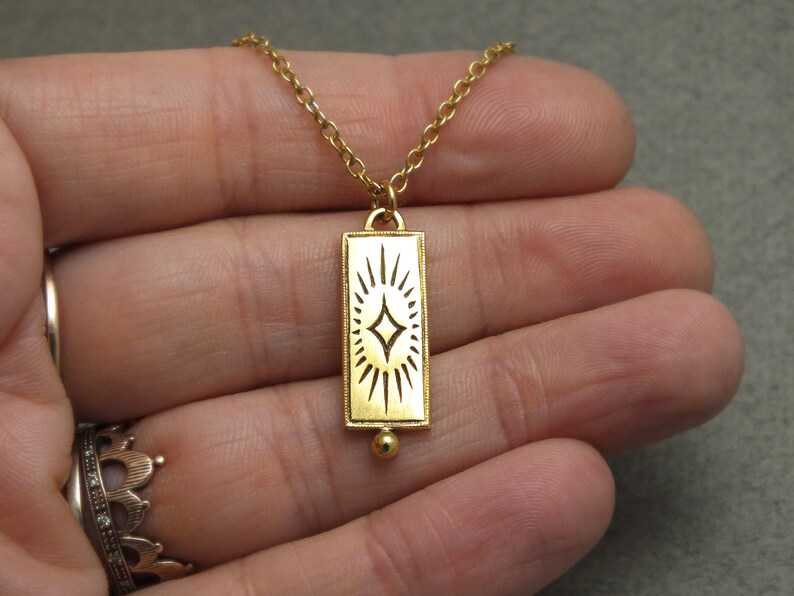 Może przedstawiać: Złoty naszyjnik z prostokątnym wisiorem w kształcie rozgwieżdżonego słońca. Wisiorek jest przymocowany do delikatnego złotego łańcuszka.