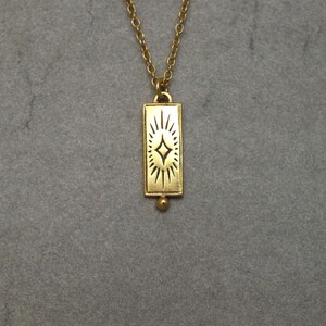 Może przedstawiać: Złoty naszyjnik z prostokątnym wisiorem w kształcie rozgwieżdżonego słońca.