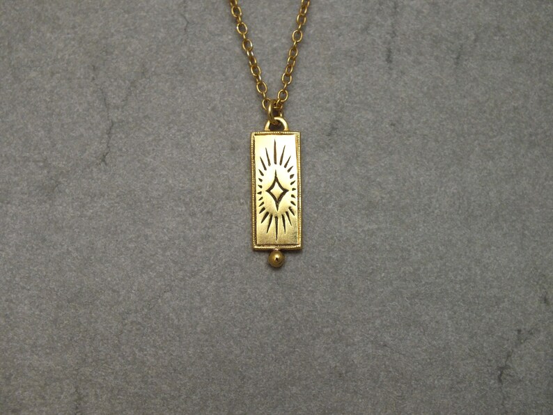 Może przedstawiać: Złoty naszyjnik z prostokątnym wisiorem w kształcie rozgwieżdżonego słońca.
