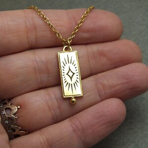 Może przedstawiać: Złoty naszyjnik z łańcuszkiem i prostokątnym wisiorem w kształcie rozgwieżdżonego słońca. Wisiorek wykonany jest ze złotego metalu i ma białą emaliowaną powierzchnię.