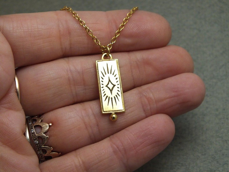 Może przedstawiać: Złoty naszyjnik z łańcuszkiem i prostokątnym wisiorem w kształcie rozgwieżdżonego słońca. Wisiorek wykonany jest ze złotego metalu i ma białą emaliowaną powierzchnię.