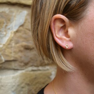 May include: A close-up of a person's ear with a small gold geometric earring. The earring is a square shape with a delicate, intricate design.