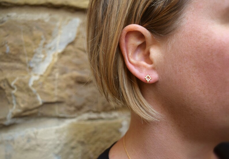May include: A close-up of a person's ear with a small gold geometric earring. The earring is a square shape with a delicate, intricate design.
