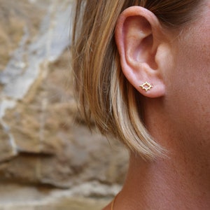 May include: A close-up of a person's ear with a small gold square earring. The earring has a geometric design with small dots around the edges.