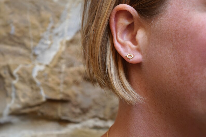 May include: A close-up of a person's ear with a small gold square earring. The earring has a geometric design with small dots around the edges.